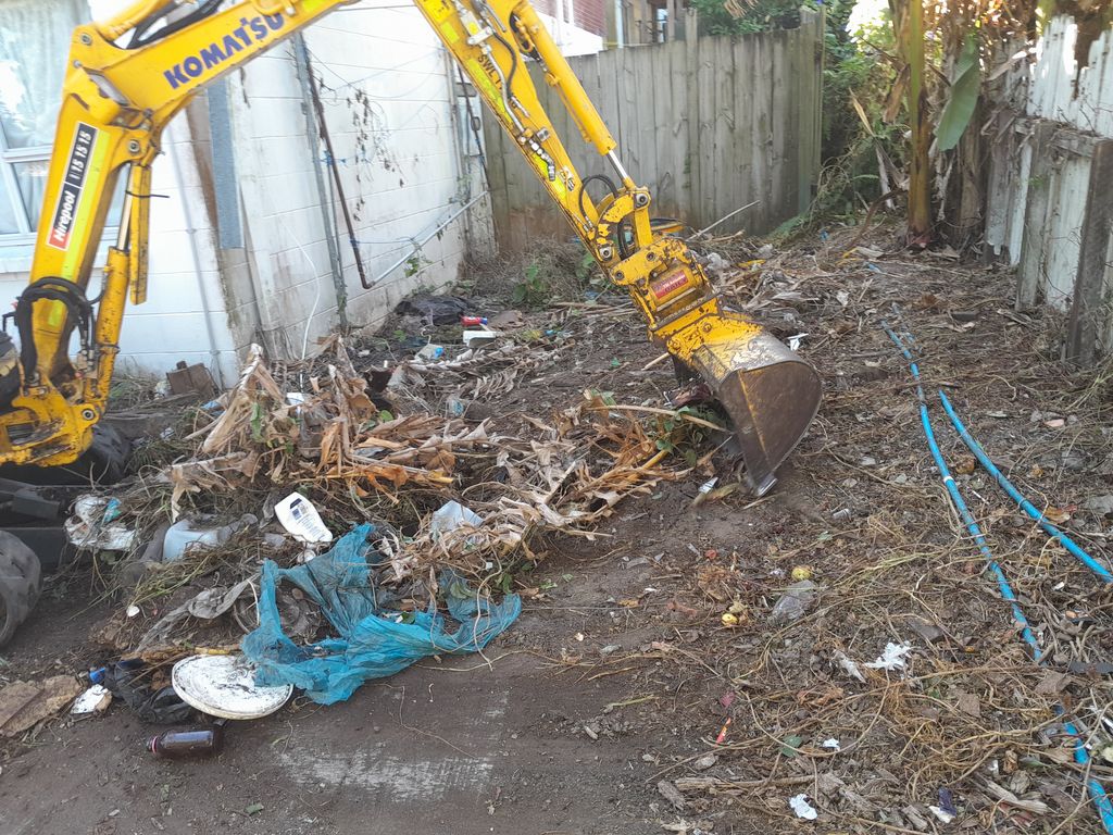 Mini digger bucket clearing branches and mixed debris beside a fence