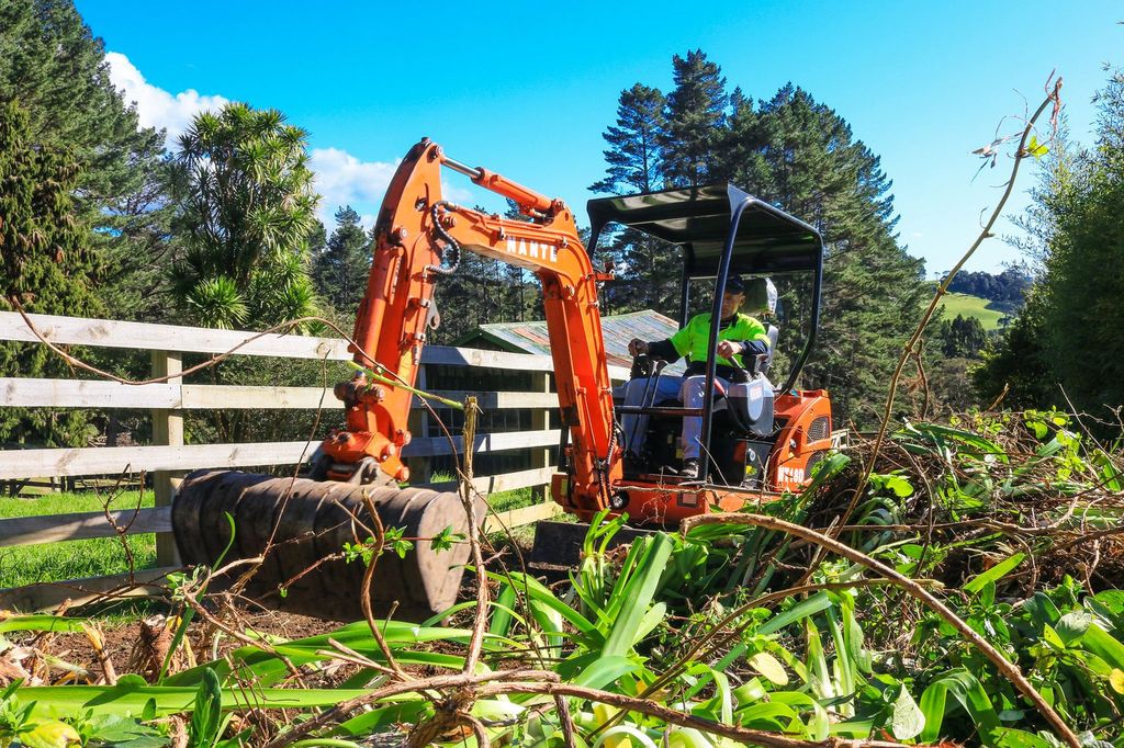 Operator clearing flax and green waste along a fence line with a mini digger